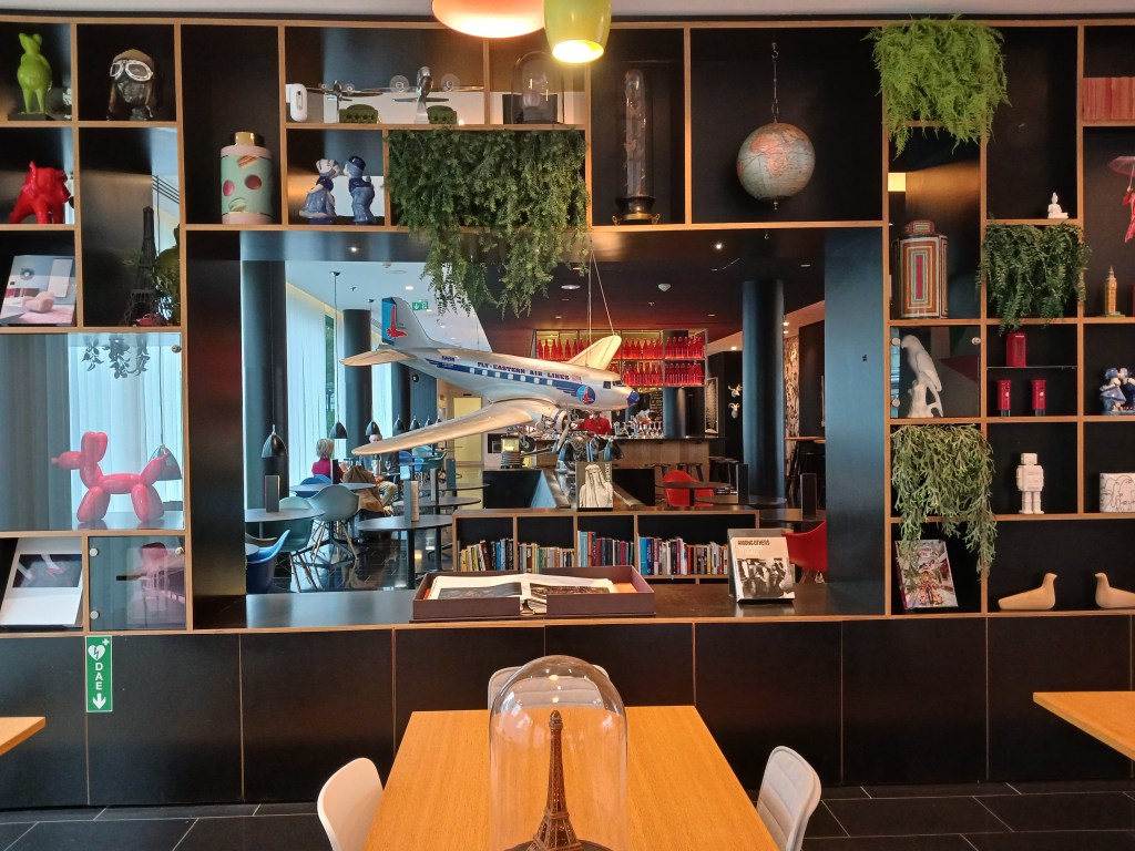 A colorful and eclectic interior design featuring a wooden shelf filled with decorative items, plants, and books. In the center, there is a model airplane hanging above a dining area with tables and chairs.