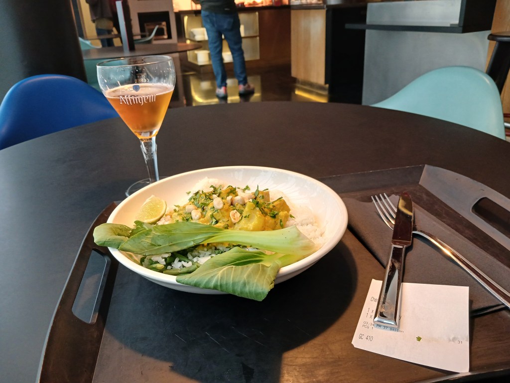 A plate of rice topped with a yellow mango dish, garnished with herbs and served with lime and greens, alongside a glass of beer on a black table.