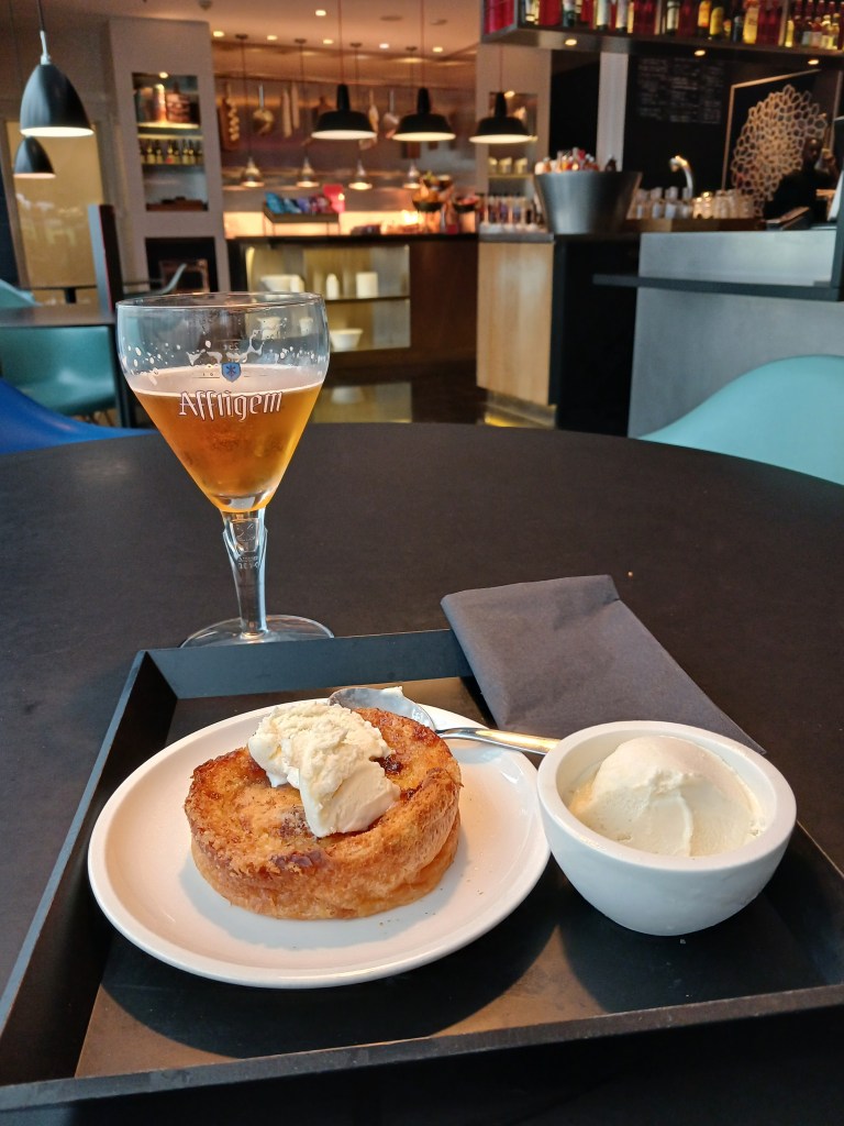 A glass of Affligem beer sits on a black table next to a dessert plate featuring a pastry topped with ice cream and a small bowl of vanilla ice cream. The background shows a stylish café with warm lighting and a counter.