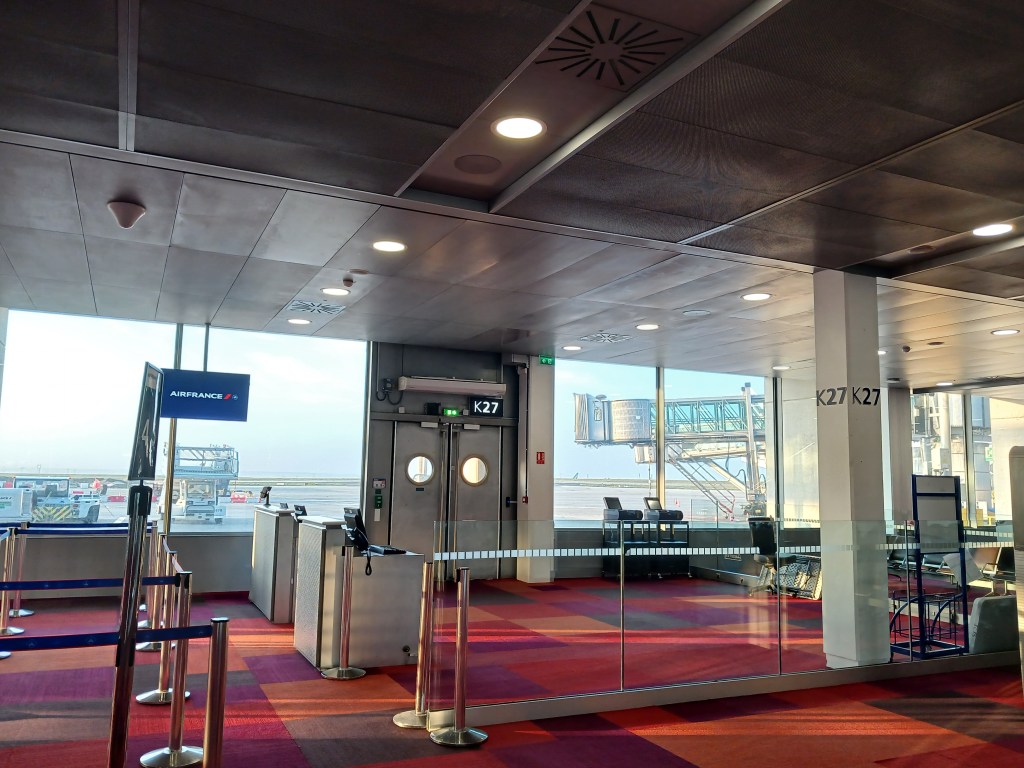 Interior view of airport terminal gate K27, featuring a departure gate with an Air France sign, seating area, and jet bridge visible through large windows.