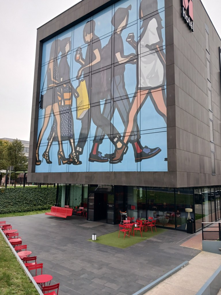 A modern hotel facade featuring a large mural of people walking, with a blue background. The ground area includes red outdoor seating in front of the entrance.