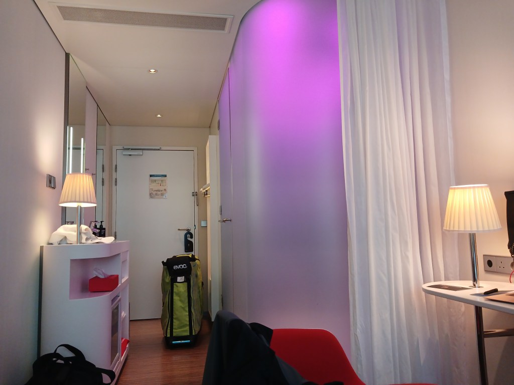 Interior view of a modern hotel room featuring a purple accent wall, a small table with a lamp, and a suitcase on the floor.
