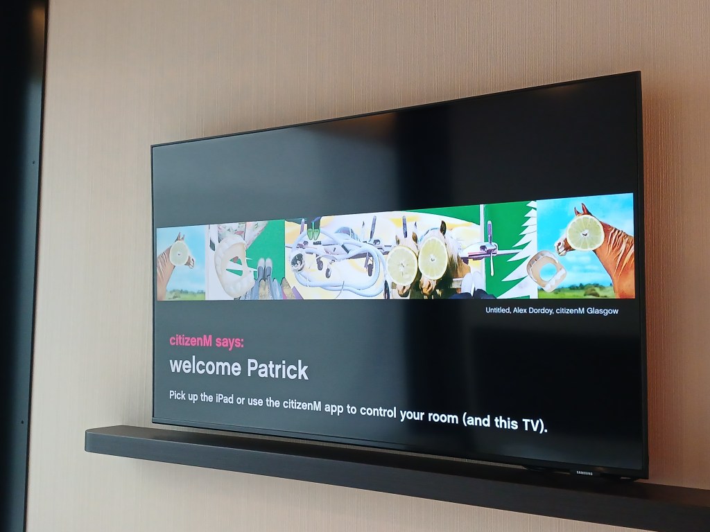 A digital screen displaying a welcome message to a guest named Patrick at citizenM, featuring colorful artwork and instructions to use an iPad for room control.