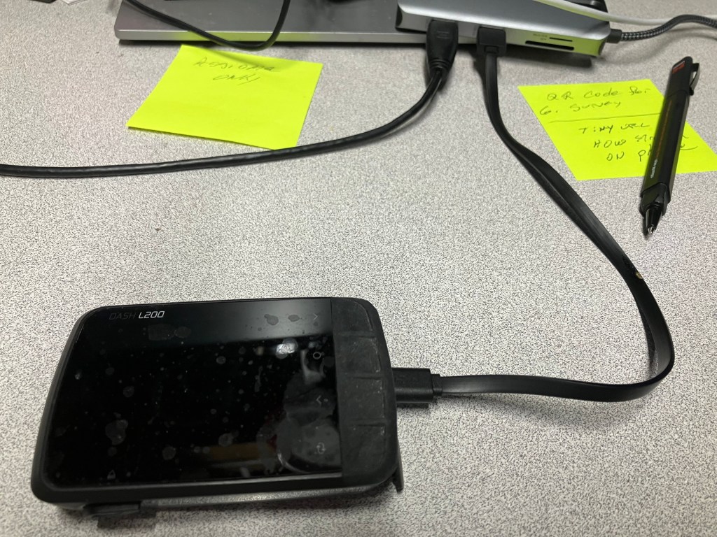 A Dash L200 device with a black screen, connected by a charging cable, situated on a desk alongside sticky notes and a pen.